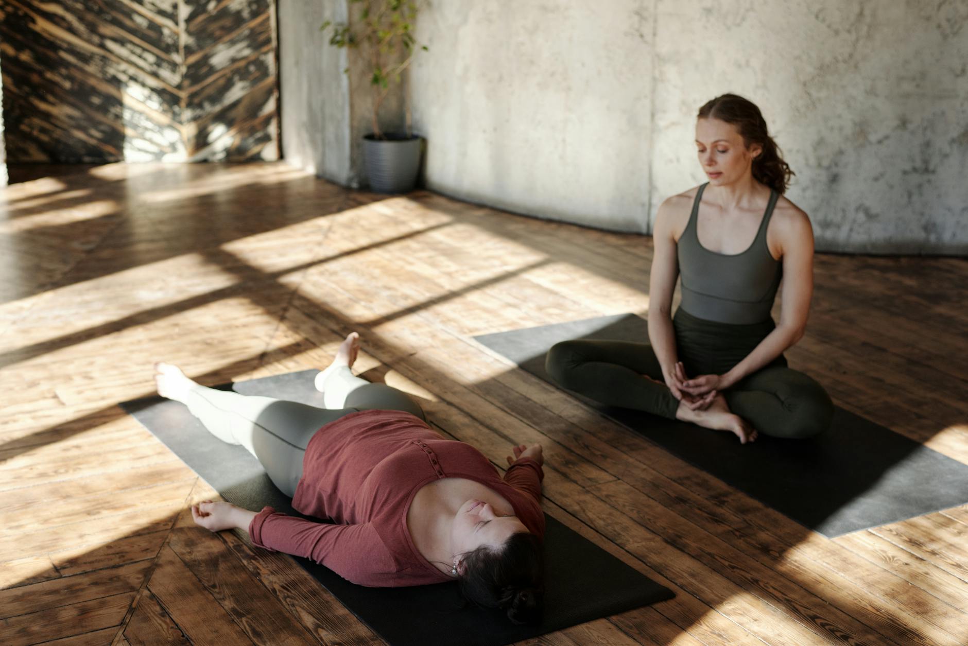 Deux femmes pratiquent des exercices de relaxation dans une salle en bois, avec une lumière naturelle traversant les fenêtres.